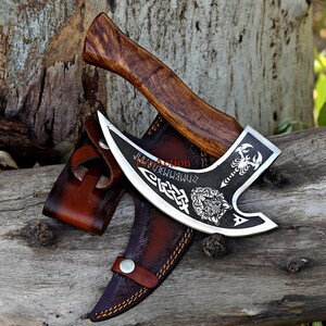 May include: A handcrafted axe with a dark wooden handle and a silver blade. The blade features intricate engravings of a scorpion, a wolf, and runes. The axe rests on a wooden surface, accompanied by a brown leather sheath.