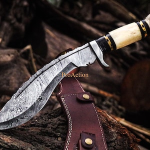 Puede incluir: Un cuchillo de acero de Damasco con una hoja curva y un mango de hueso. El cuchillo tiene una funda de cuero con remaches de color lat&oacute;n. La hoja tiene un patr&oacute;n &uacute;nico y el mango est&aacute; acentuado con detalles negros y dorados.