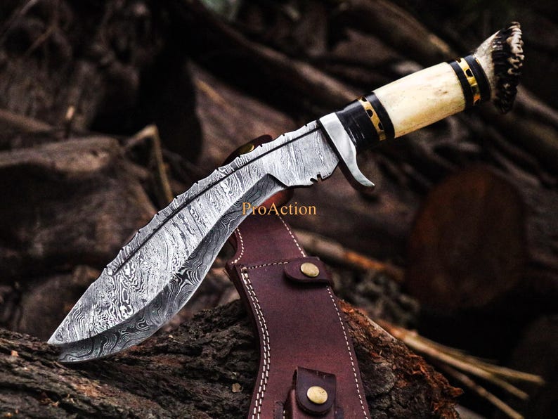 Puede incluir: Un cuchillo de acero de Damasco con una hoja curva y un mango de hueso. El cuchillo tiene una funda de cuero con remaches de color lat&oacute;n. La hoja tiene un patr&oacute;n &uacute;nico y el mango est&aacute; acentuado con detalles negros y dorados.