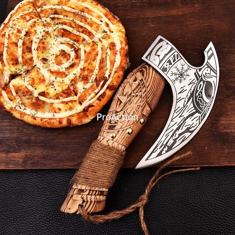 Op de afbeelding: Een decoratieve bijl met een houten handvat en een zilveren blad met gegraveerde ontwerpen. De handgreep is omwikkeld met touw. Op de achtergrond een pizza met een spiraal van witte saus.