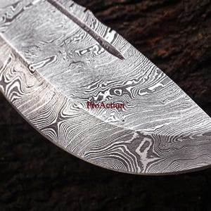 May include: Close-up of a Damascus steel knife blade. The blade has a distinctive pattern of swirling lines in shades of gray and white. The text "ProAction" is visible on the blade. The knife blade is curved and has a sharp point.