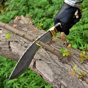 Puede incluir: Un cuchillo grande con una hoja oscura y un mango de lat&oacute;n y negro est&aacute; cortando una rama de &aacute;rbol. El cuchillo tiene una hoja texturizada y lo sostiene una mano enguantada. El fondo es una vista borrosa de follaje verde.