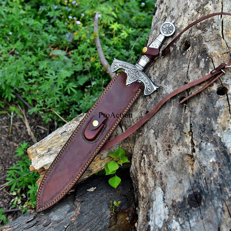 Puede incluir: Una espada plateada con una empu&ntilde;adura decorativa y una funda de cuero marr&oacute;n. La funda est&aacute; cosida con hilo dorado y tiene una correa de cuero. La espada descansa sobre una superficie de madera envejecida.