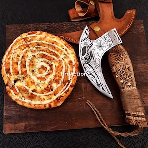 Op de afbeelding: Een decoratieve Vikingbijl met een houten handvat en een leren schede ligt op een houten snijplank naast een pizza. Het bijlblad heeft ingewikkelde gravures. De pizza heeft een spiraalvormig patroon.