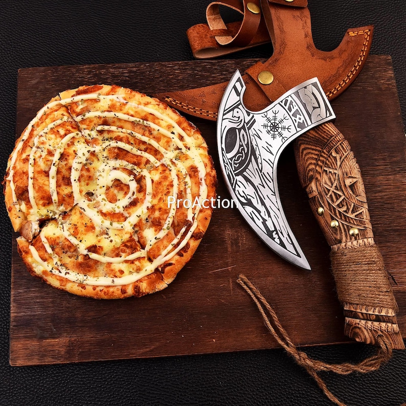 Op de afbeelding: Een decoratieve Vikingbijl met een houten handvat en een leren schede ligt op een houten snijplank naast een pizza. Het bijlblad heeft ingewikkelde gravures. De pizza heeft een spiraalvormig patroon.
