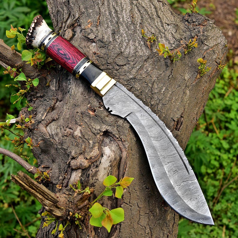 Puede incluir: Un cuchillo grande y curvo con una hoja de acero estampada y un mango de madera rojo y negro. El mango tiene detalles de lat&oacute;n y un extremo de asta blanco y marr&oacute;n. El cuchillo est&aacute; apoyado en un tronco de &aacute;rbol.