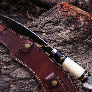 Puede incluir: Un cuchillo hecho a mano con una hoja negra y un mango de hueso, en una funda de cuero marr&oacute;n. El cuchillo tiene un borde dentado y est&aacute; acentuado con detalles en negro y dorado. La funda est&aacute; asegurada con tachuelas color lat&oacute;n.