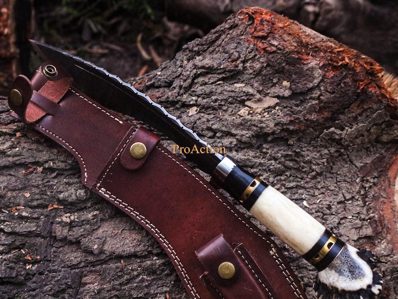 Puede incluir: Un cuchillo hecho a mano con una hoja negra y un mango de hueso, en una funda de cuero marr&oacute;n. El cuchillo tiene un borde dentado y est&aacute; acentuado con detalles en negro y dorado. La funda est&aacute; asegurada con tachuelas color lat&oacute;n.