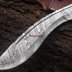 May include: Close-up of a Damascus steel knife blade with a curved edge. The blade has a unique, swirling pattern in shades of silver and black. The text "ProAction" is visible on the blade. The knife is likely for hunting or outdoor use.
