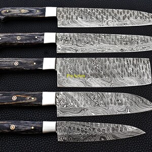 Puede incluir: Un juego de cinco cuchillos de cocina de acero de Damasco con mangos de madera oscura. Las hojas tienen un patr&oacute;n arremolinado &uacute;nico. Cada cuchillo tiene una virola plateada y remaches de lat&oacute;n en el mango. Los cuchillos est&aacute;n sobre una superficie negra.