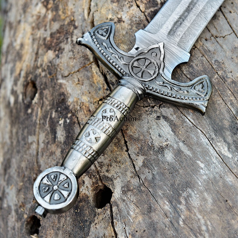 Puede incluir: Primer plano de una espada plateada con un mango ornamentado y dise&ntilde;os en forma de cruz. La hoja tiene una textura estampada. La espada descansa sobre una superficie de madera envejecida, mostrando los detalles del arma de estilo medieval.