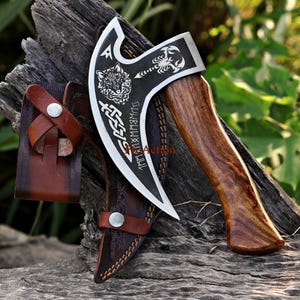 May include: A handcrafted axe with a dark blade featuring a wolf, scorpion, and runic symbols. The axe has a wooden handle and comes with a brown leather sheath. The blade is approximately 6 inches long.