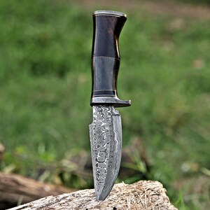 Pode incluir: Uma faca de ca&ccedil;a artesanal com cabo de madeira escura e l&acirc;mina de a&ccedil;o estampada. A l&acirc;mina tem um design &uacute;nico em espiral. A faca est&aacute; apoiada em um peda&ccedil;o de madeira contra um fundo verde desfocado.