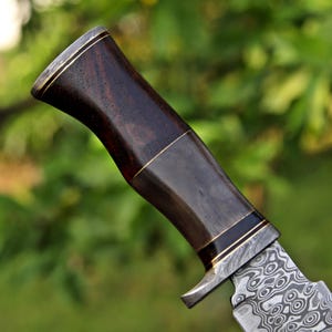 Pode incluir: Close-up de uma faca com cabo de madeira escura e l&acirc;mina de a&ccedil;o estampada. A pega apresenta detalhes em lat&atilde;o e um design curvo. A l&acirc;mina tem um padr&atilde;o de redemoinho &uacute;nico.