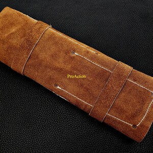 Puede incluir: Un estuche rectangular de cuero de ante marr&oacute;n con costuras blancas, dos correas de cuero y cuatro remaches met&aacute;licos. La palabra ProAction est&aacute; impresa en amarillo en el centro de la superficie exterior del estuche de cuero marr&oacute;n.