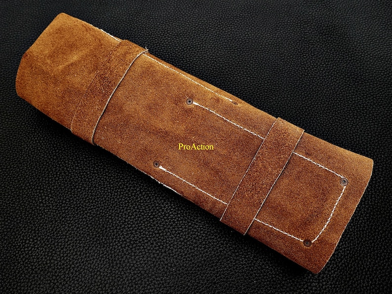 Puede incluir: Un estuche rectangular de cuero de ante marr&oacute;n con costuras blancas, dos correas de cuero y cuatro remaches met&aacute;licos. La palabra ProAction est&aacute; impresa en amarillo en el centro de la superficie exterior del estuche de cuero marr&oacute;n.