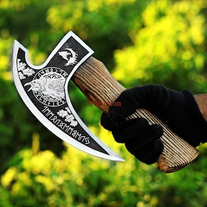 May include: A silver and black axe with a wooden handle. The axe head features engraved imagery of a raven, wolf, and runes. The handle is made of wood and has a textured appearance. The axe is held by a person wearing a black glove.