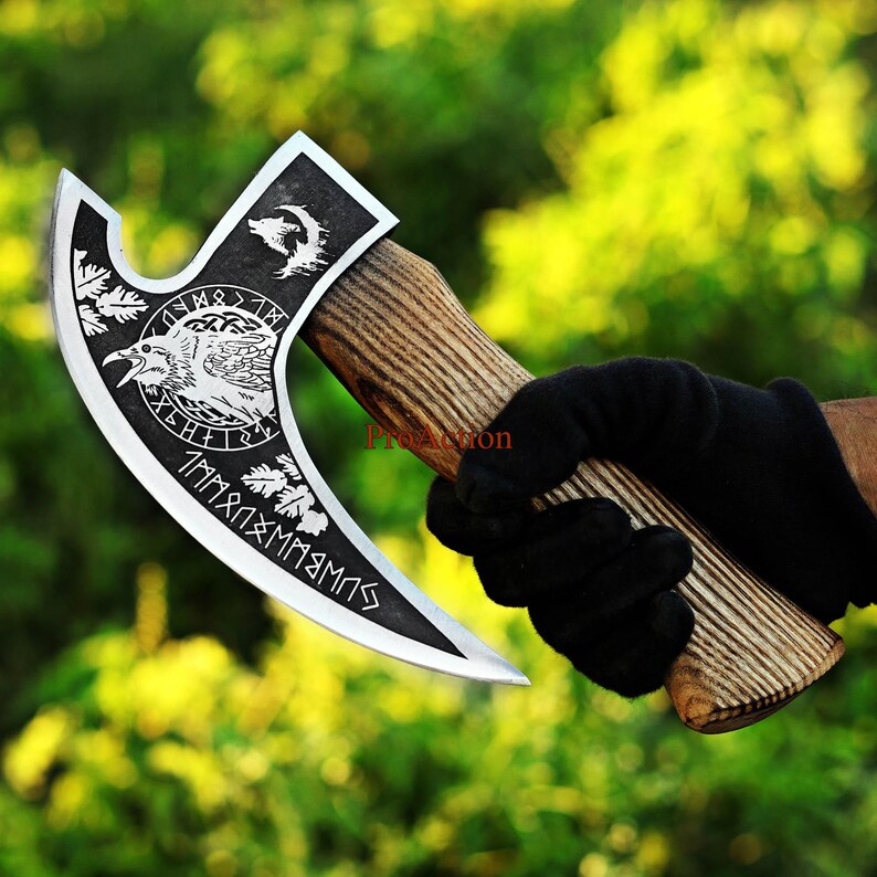 May include: A silver and black axe with a wooden handle. The axe head features engraved imagery of a raven, wolf, and runes. The handle is made of wood and has a textured appearance. The axe is held by a person wearing a black glove.