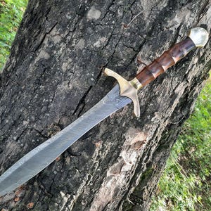 May include: A decorative sword with a patterned blade and a wooden handle. The handle is dark brown with a brass guard and pommel. The sword is resting on a tree trunk.