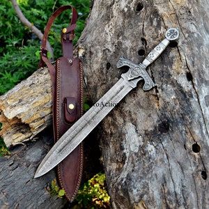 Puede incluir: Una espada plateada con una empu&ntilde;adura ornamentada y una hoja texturizada descansa sobre una superficie de madera envejecida. Una funda de cuero marr&oacute;n con una correa para el hombro est&aacute; a la izquierda. La espada presenta un dise&ntilde;o de cruz en la empu&ntilde;adura y la guarda.