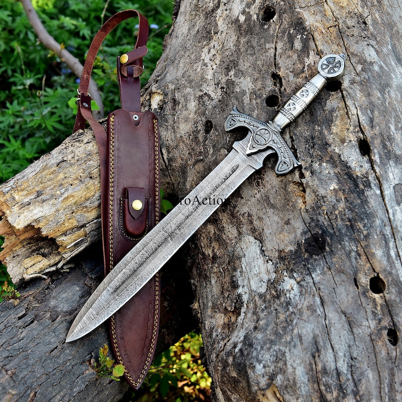 Puede incluir: Una espada plateada con una empu&ntilde;adura ornamentada y una hoja texturizada descansa sobre una superficie de madera envejecida. Una funda de cuero marr&oacute;n con una correa para el hombro est&aacute; a la izquierda. La espada presenta un dise&ntilde;o de cruz en la empu&ntilde;adura y la guarda.