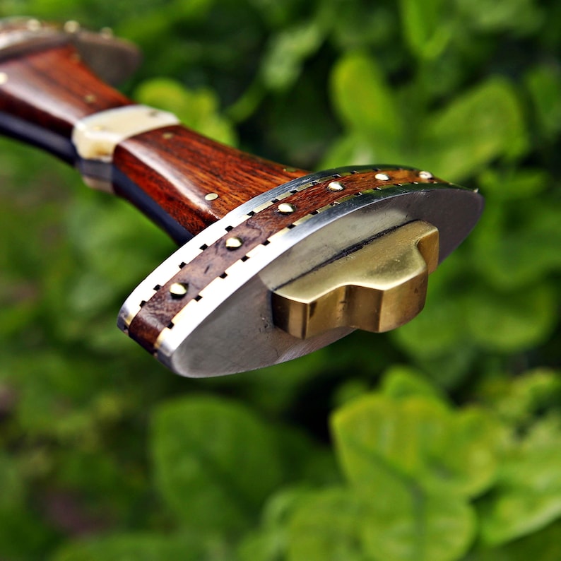 May include: Close-up of a handcrafted knife handle. The handle features dark wood, brass accents, and a polished metal bolster. Small brass rivets secure the handle's components. The blade is not visible.