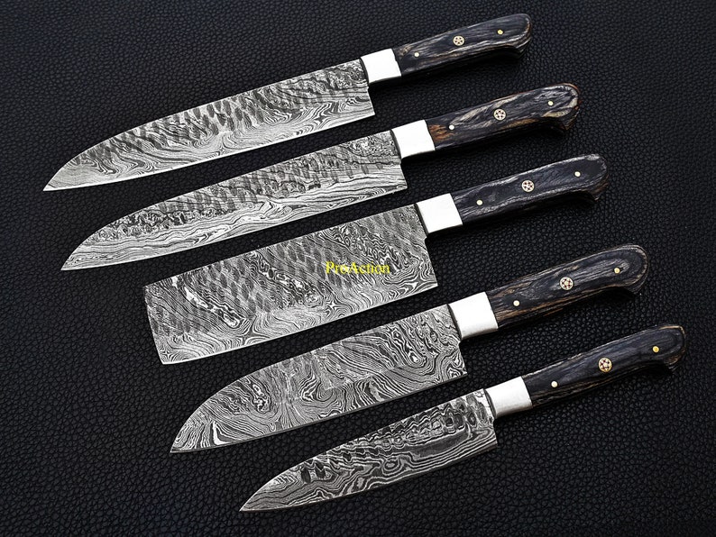 Puede incluir: Juego de cinco cuchillos de cocina de acero de Damasco con mangos de madera oscura. Las hojas tienen un dise&ntilde;o distintivo. Cada cuchillo tiene una virola plateada y remaches de lat&oacute;n. Dispuestos sobre una superficie negra.