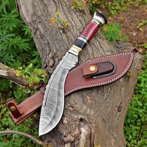 Puede incluir: Un cuchillo grande y curvo con una hoja estampada y un mango rojo y negro. El cuchillo descansa sobre un tronco de &aacute;rbol junto a una funda de cuero marr&oacute;n. El mango presenta detalles en lat&oacute;n y una secci&oacute;n de asta.