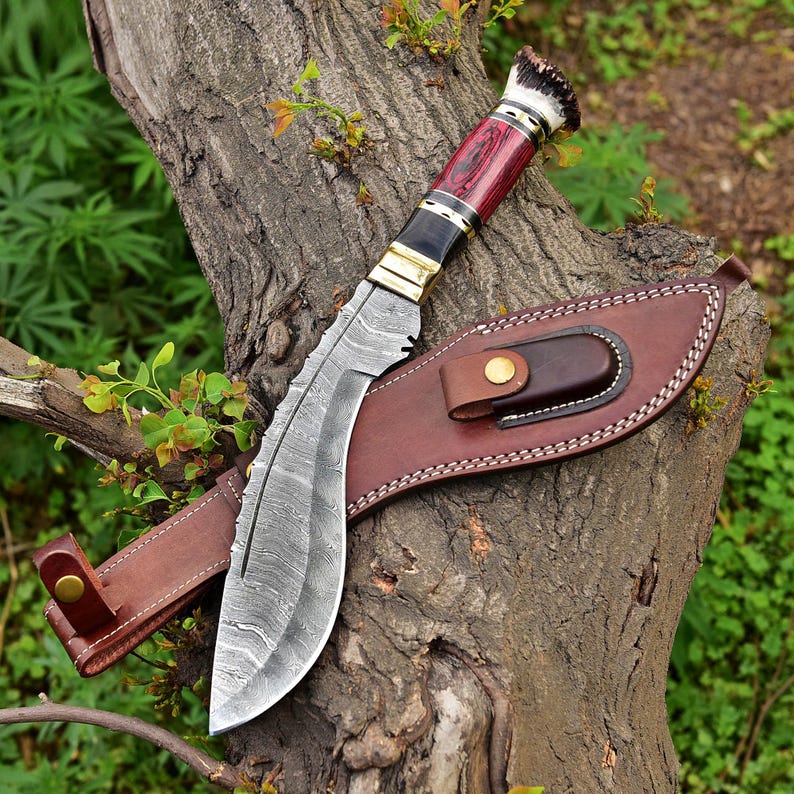 Puede incluir: Un cuchillo grande y curvo con una hoja estampada y un mango rojo y negro. El cuchillo descansa sobre un tronco de &aacute;rbol junto a una funda de cuero marr&oacute;n. El mango presenta detalles en lat&oacute;n y una secci&oacute;n de asta.