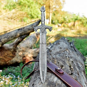 Puede incluir: Una espada plateada con una empu&ntilde;adura ornamentada y una hoja estampada. La espada se muestra de pie sobre un tronco desgastado, con una funda de cuero marr&oacute;n. El texto "ProAction" es visible en la hoja.
