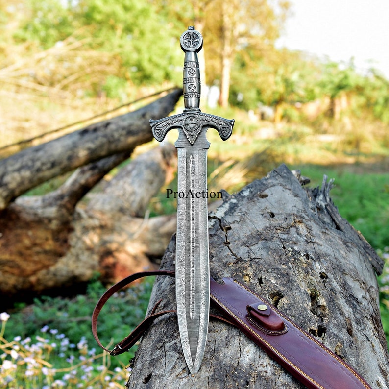 Puede incluir: Una espada plateada con una empu&ntilde;adura ornamentada y una hoja estampada. La espada se muestra de pie sobre un tronco desgastado, con una funda de cuero marr&oacute;n. El texto "ProAction" es visible en la hoja.