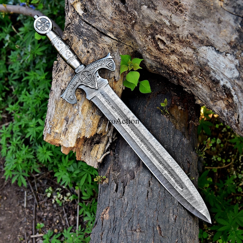 Puede incluir: Una espada plateada con un mango ornamentado y un dise&ntilde;o en forma de cruz. La hoja tiene una textura estampada. La espada descansa sobre una superficie de madera envejecida, con follaje verde de fondo.