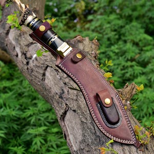 Puede incluir: Un cuchillo artesanal con un mango oscuro y una funda de cuero marr&oacute;n. El cuchillo presenta un mango decorativo con detalles dorados y una hoja. La funda tiene un lazo para cintur&oacute;n y se asegura con un broche.