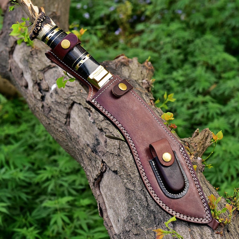 Puede incluir: Un cuchillo artesanal con un mango oscuro y una funda de cuero marr&oacute;n. El cuchillo presenta un mango decorativo con detalles dorados y una hoja. La funda tiene un lazo para cintur&oacute;n y se asegura con un broche.