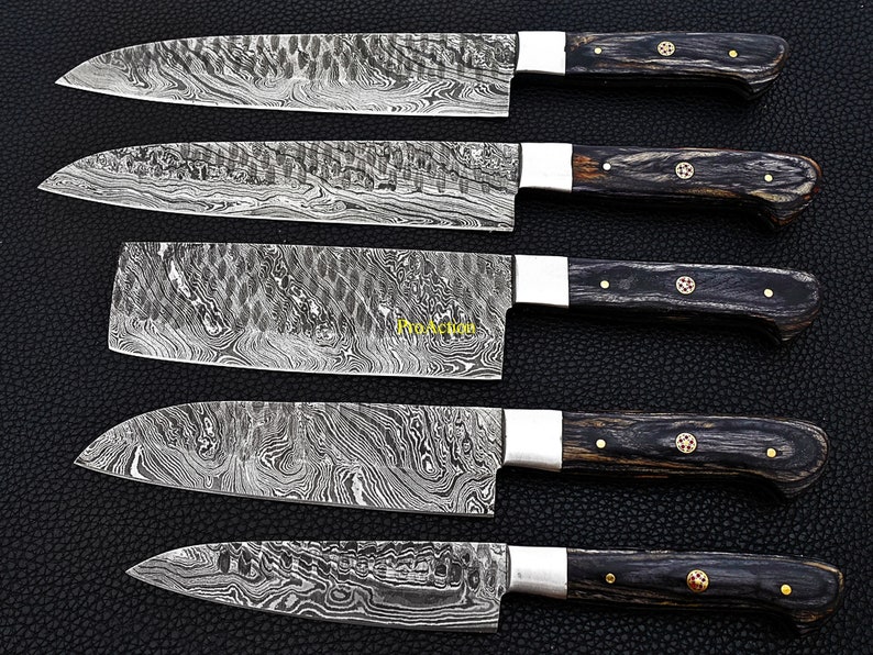 Puede incluir: Un surtido de cinco cuchillos de cocina de acero de Damasco. Las hojas tienen un dise&ntilde;o &uacute;nico y estampado. Cada cuchillo presenta un mango de madera oscura, virolas plateadas y remaches decorativos. Los cuchillos est&aacute;n dispuestos sobre una superficie oscura.