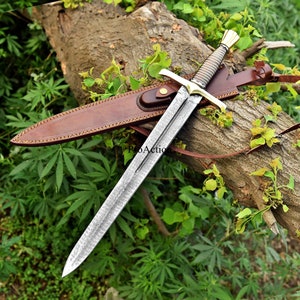 May include: A long, ornate sword with a patterned blade and gold-colored accents, resting on a log with a brown leather sheath. The sword features a detailed handle and crossguard. The background is lush green foliage.