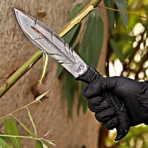 Puede incluir: Un cuchillo de acero de Damasco con un dise&ntilde;o en forma de pluma. La hoja tiene un patr&oacute;n en capas con detalles dorados. El mango es negro y texturizado. El cuchillo es sostenido por una mano enguantada sobre un fondo verde borroso.
