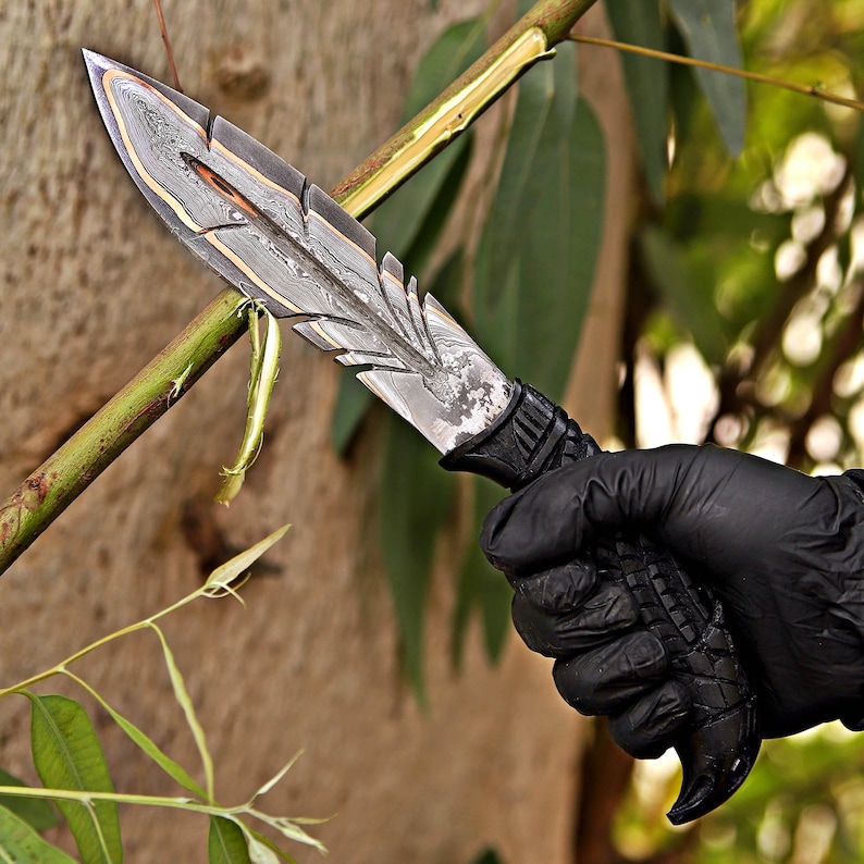 Puede incluir: Un cuchillo de acero de Damasco con un dise&ntilde;o en forma de pluma. La hoja tiene un patr&oacute;n en capas con detalles dorados. El mango es negro y texturizado. El cuchillo es sostenido por una mano enguantada sobre un fondo verde borroso.
