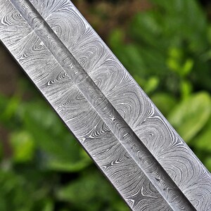 May include: Close-up of a sword blade with a distinctive pattern. The blade features a swirling, wave-like design in shades of gray and silver, creating a textured appearance. The central ridge of the blade is visible, with a blurred green background.
