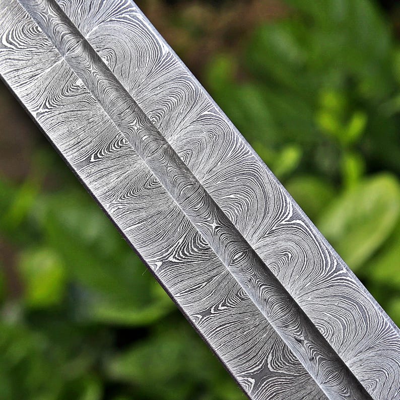 May include: Close-up of a sword blade with a distinctive pattern. The blade features a swirling, wave-like design in shades of gray and silver, creating a textured appearance. The central ridge of the blade is visible, with a blurred green background.