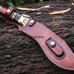 May include: A handcrafted knife with a brown leather sheath. The knife handle features red, black, and gold accents, with a decorative end. The sheath is stitched with white thread and secured with brass-colored rivets.