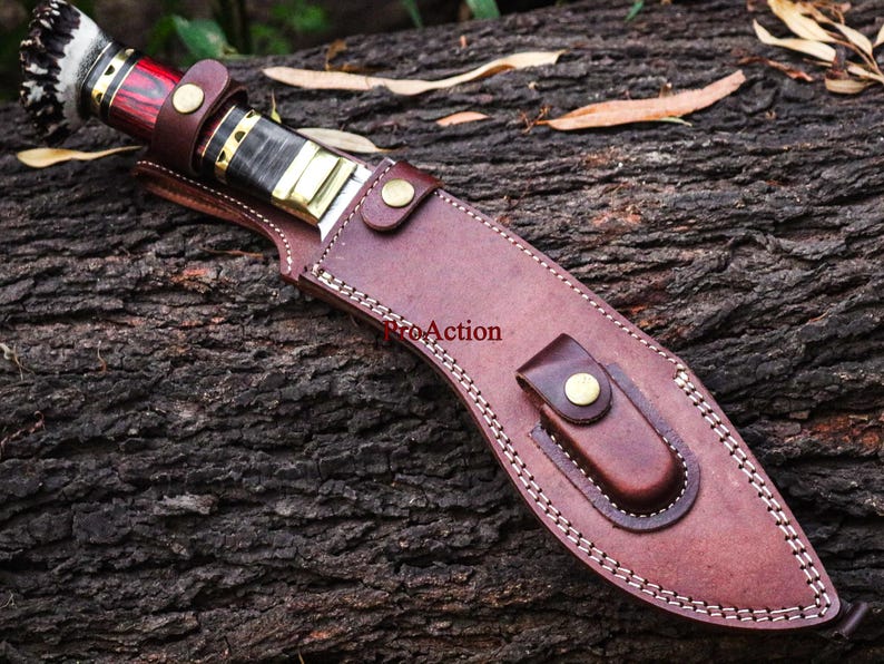 May include: A handcrafted knife with a brown leather sheath. The knife handle features red, black, and gold accents, with a decorative end. The sheath is stitched with white thread and secured with brass-colored rivets.