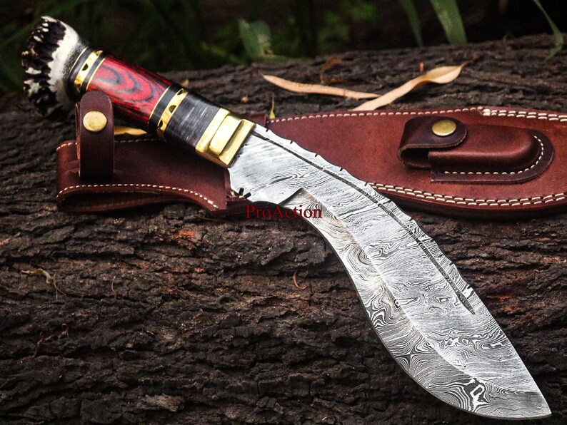 May include: A Damascus steel kukri knife with a red and black wooden handle, brass accents, and a leather sheath. The blade has a distinctive curved shape and a patterned surface. The knife is designed for outdoor use.
