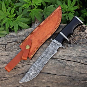 May include: A Damascus steel hunting knife with a black handle and a brown leather sheath. The blade has a distinctive pattern, and the sheath is secured with a brass button. The knife is displayed on a wooden surface.