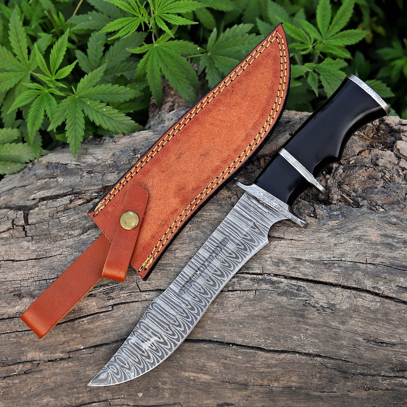May include: A Damascus steel hunting knife with a black handle and a brown leather sheath. The blade has a distinctive pattern, and the sheath is secured with a brass button. The knife is displayed on a wooden surface.