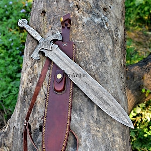 Puede incluir: Espada decorativa con una hoja estampada y un mango ornamentado, enfundada en una vaina de cuero marr&oacute;n. La vaina tiene una correa para el hombro. La espada y la vaina est&aacute;n sobre un tronco de &aacute;rbol.