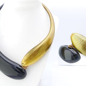 Könnte beinhalten: Ein modernes Halsketten- und Ohrring-Set. Die Halskette hat einen schwarzen, gebogenen Anhänger, der mit einem goldfarbenen, strukturierten Band verbunden ist. Die Ohrringe haben ein ähnliches Design, mit einem schwarzen Stein und einem goldfarbenen, strukturierten Oval. Schmuck.