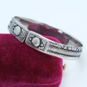 Puede incluir: Un brazalete de plata con un intrincado diseño floral y dos acentos circulares. El brazalete está abierto y tiene un cierre con bisagra.