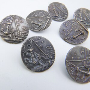 May include: Seven silver-toned metal buttons with an embossed design of a sailboat and anchor. The buttons are round and have a raised edge.