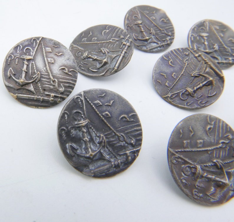 May include: Seven silver-toned metal buttons with an embossed design of a sailboat and anchor. The buttons are round and have a raised edge.